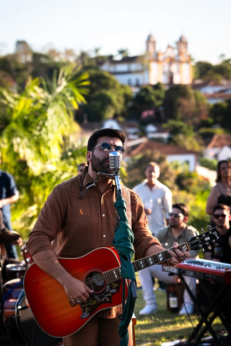 Geovani Medeiros sentado em uma banqueta tocando violão, cercado pelos convidados no gramado